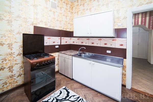 Компактная кухня с белыми шкафами, плитой, мойкой, цветочными обоями и ковриком с чёрно‑белым узором.