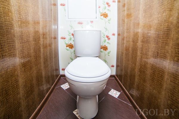 Небольшой санузел с белым унитазом, цветочными обоями и тёмным деревянным полом.