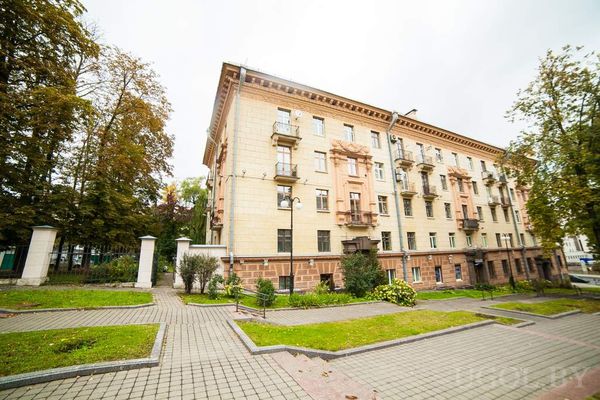 Многоэтажный жилой дом с балконами и красными акцентами, во дворе зелёная зона с лавочками и дорожками.