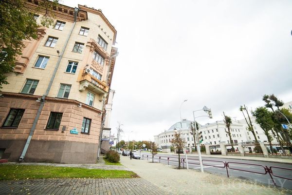 Городская улица с многоэтажным домом слева и классическими зданиями справа, пешеходами и деревьями вдоль дороги.