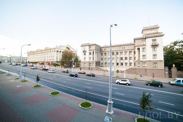 Широкая городская улица с машинами, автобусом и зданием с колоннами и треугольным фронтоном.