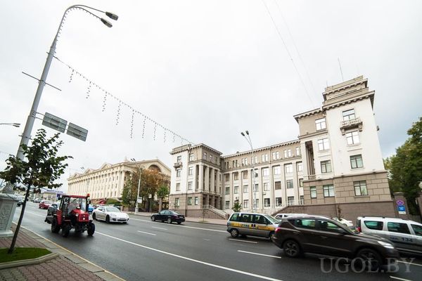 Городская улица с машинами и красным трактором, зданиями с колоннами и облачным небом.