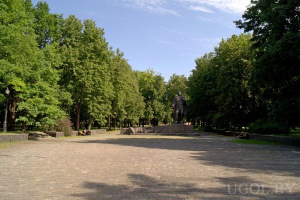 Площадь, вымощенная плиткой, с памятником на постаменте в центре и аллеей высоких зелёных деревьев по бокам.