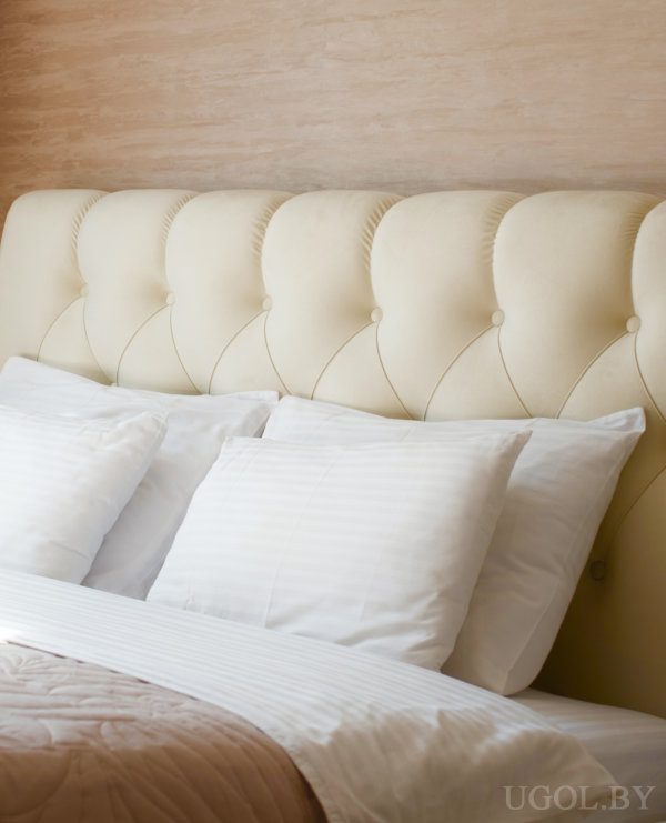 Крупный план белой постели с кремовым изголовьем в ромб, на фоне тёплой стены и текстиля.