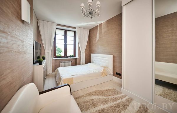 Белая кровать, диван, ТВ и шкаф-купе с зеркалом, интерьер в светлых тонах с люстрой и растениями.