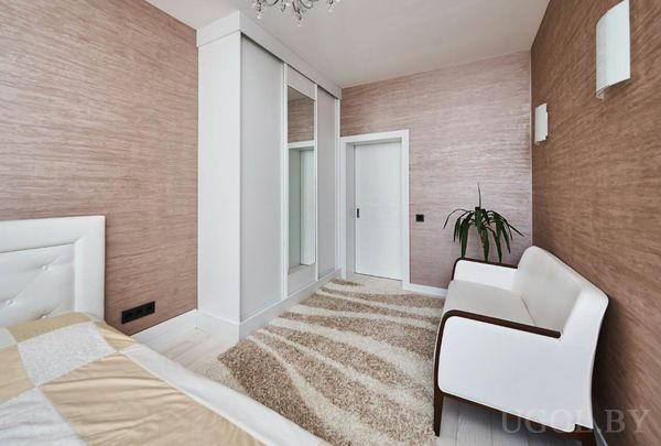 Белая кровать, кресло с растением, шкаф с зеркалом и тёплая отделка стен, подчёркивающая уют.