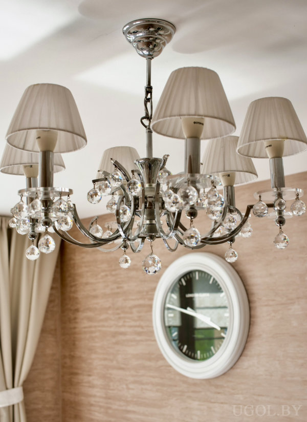 Классическая люстра с подвесками и настенные часы на фоне бежевых штор и текстурной стены.