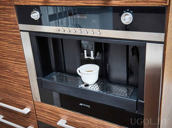 Чашка кофе на поддоне, кнопки и регуляторы на панели, техника встроена в деревянный фасад.