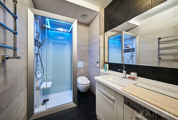 Душ с синей подсветкой, белая тумба, зеркало и полотенцесушитель на фоне светло-тёмной плитки.