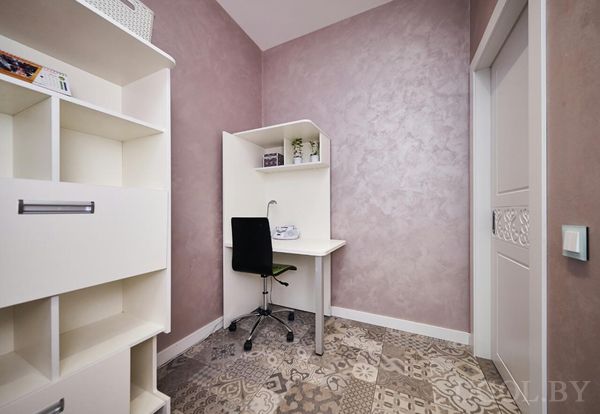 Белый стол, принтер, чёрное кресло и полки с декором на фоне сиреневой стены и узорного пола.