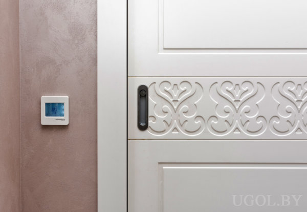 Белая раздвижная дверь с узором, термостат на стене и тёплая отделка в современном интерьере.