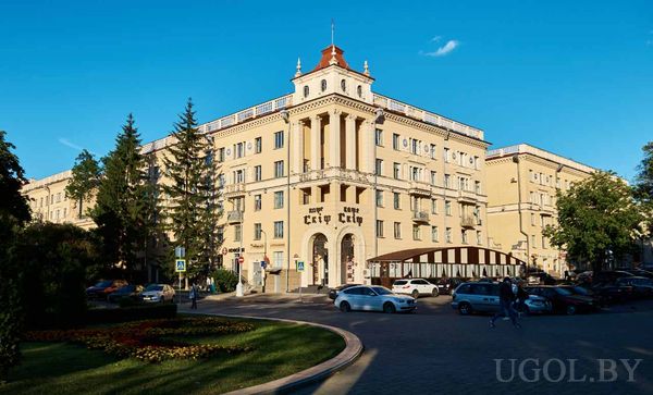 Многоэтажное здание с колоннами, башней и вывеской «City City», перед ним авто, люди и зелёные насаждения.