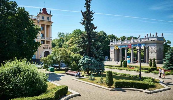 Прогулочная зона с лавочками, деревьями и аркой с колоннами, рядом здание с красной крышей и вывеской «City».