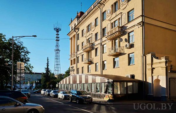 Бежевое здание с балконами и стеклянной пристройкой, рядом телекоммуникационная вышка и припаркованные машины.