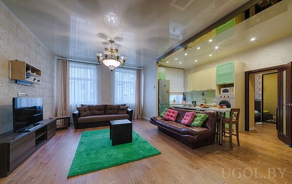 Современное пространство с двумя диванами, зелёным ковром, ТВ-зоной и кухней с мятными шкафами.