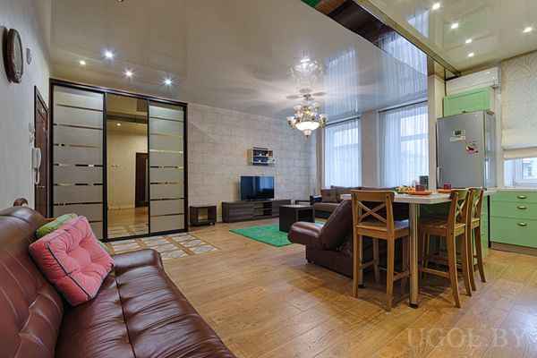 Открытая планировка с кожаным диваном, деревянным столом, мятной кухней и глянцевым потолком с люстрой.