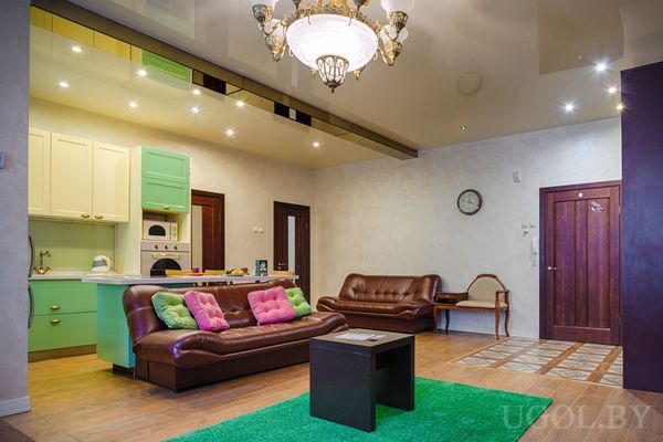 Интерьер с яркими подушками, зелёным ковром, пастельной кухней и декоративной люстрой.