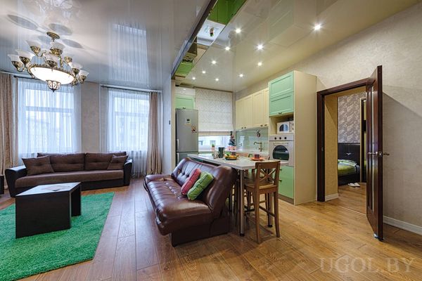 Два дивана, зелёный ковёр, кухня с барными стульями, глянцевый потолок и открытая дверь в спальню с зелёным покрывалом.