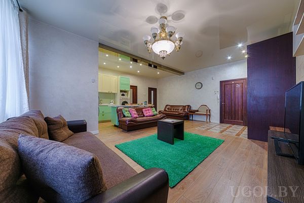 Большой диван, зелёный ковёр, мятная кухня, настенные часы и небольшой стол с креслом.