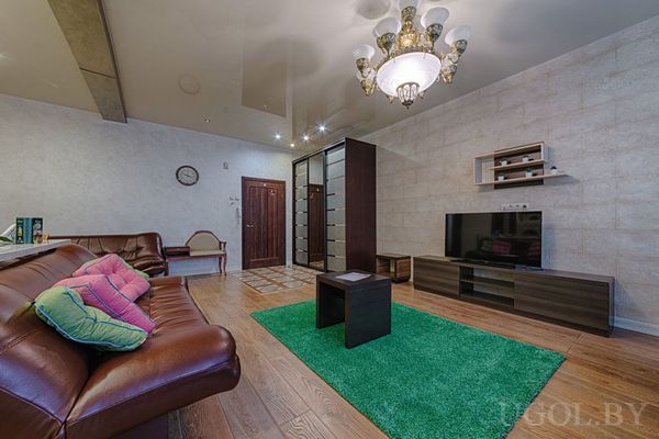 Кожаный диван с яркими подушками, ТВ-зона, зелёный ковёр, люстра и деревянная дверь с часами.