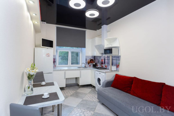 Белая кухня с техникой, стол с сервировкой, серый диван с красными подушками и потолок с круглыми светильниками.