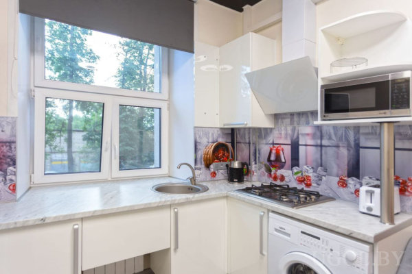 Белая мебель, газовая плита, микроволновка, стиральная машина и фартук с изображением банок и яблок.