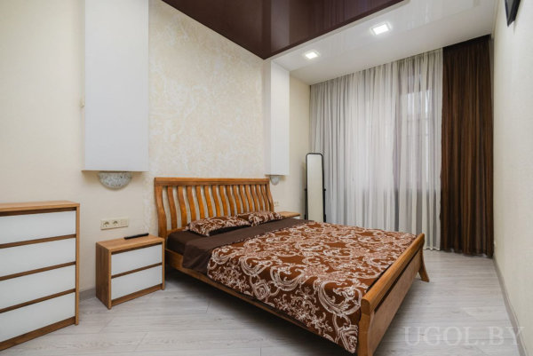 Спальня с коричневым глянцевым потолком, светлой мебелью и лаконичным дизайном.