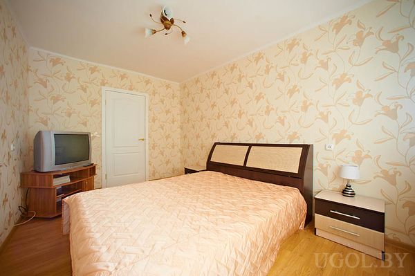 Светлая комната с цветочными обоями, тумба с лампой, CRT-телевизор, дверь на кухню, акцент на чистоту и винтажные детали.