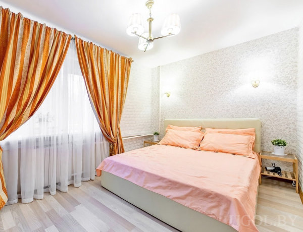 Комната с кроватью, прикроватными тумбами, растениями, настенными светильниками и шторами с золотыми полосами.