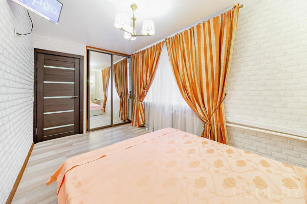 Интерьер с кроватью, зеркальным шкафом, телевизором, белой кирпичной стеной и тёмной дверью.