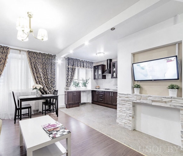 Комната с камином, телевизором, журнальным столиком, обеденной зоной и кухней с тёмными фасадами.