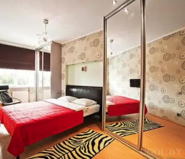 Стильная спальня с черной спинкой кровати, красным покрывалом и белыми подушками. Зебровые коврики, зеркальный шкаф и декоративная люстра создают атмосферу уюта и современности.