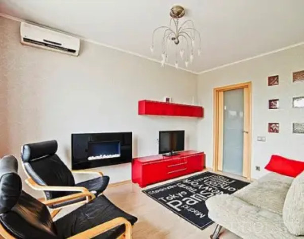 Гостиная с настенным кондиционером, черным электрокамином, красной ТВ-тумбой и подвесной люстрой. Интерьер дополнен черными креслами, светлым диваном и ковром с текстом.