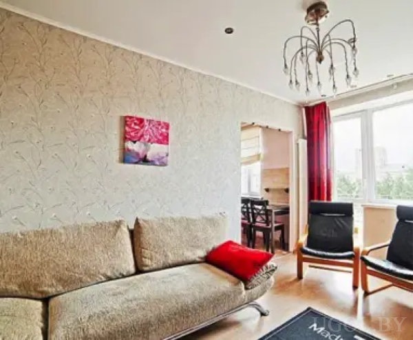 Бежевый диван, черные кресла и красные шторы у окна. На стене — картина с цветами, а за аркой — столовая зона. Интерьер оформлен в теплых тонах.