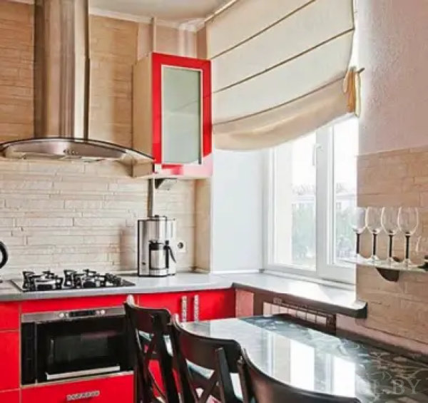 Компактная кухня с красными фасадами, кофеваркой, стеклянным столом и настенным держателем для бокалов. Светлая плитка и римская штора создают уютную атмосферу.