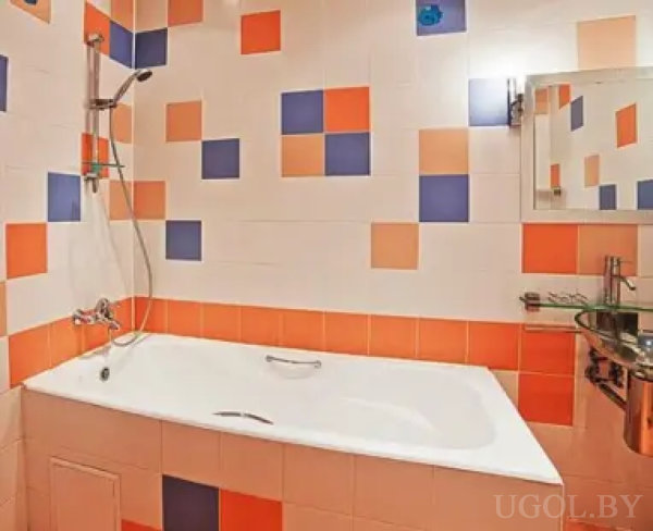 Интерьер ванной с белой ванной, душем и мозаичной плиткой в оранжево-синих тонах. Зеркало с полочкой и стеклянная полка дополняют функциональность пространства.