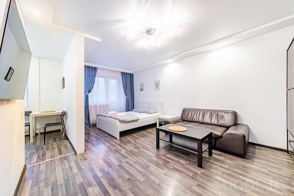 Комната с кроватью, кожаным диваном, столом, телевизором на стене и окнами с синими шторами.