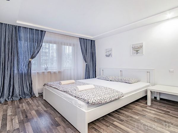 Интерьер с кроватью, прикроватной тумбой, картинами, синими шторами и тёмным ламинатом.