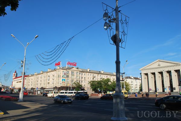 Уличная сцена с машинами, домом с рекламой, колоннами, фонарями и голубым небом.