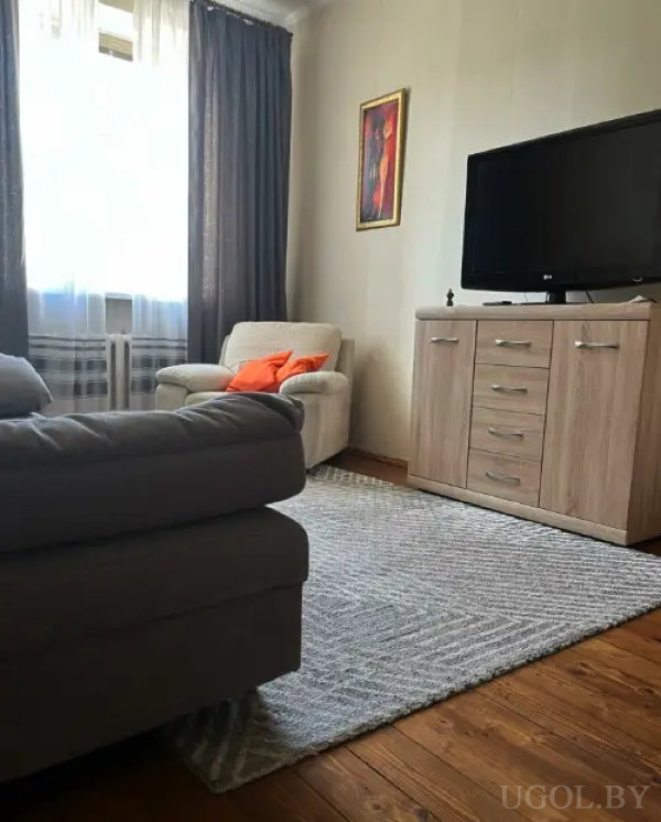 Комфортная гостиная с бежевым креслом и яркими подушками, серым диваном, телевизором на деревянной тумбе и серым ковром.