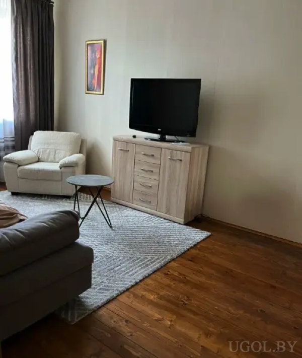 Интерьер с бежевым креслом у окна, черным круглым столиком, серым диваном и ковром с геометрическим узором.