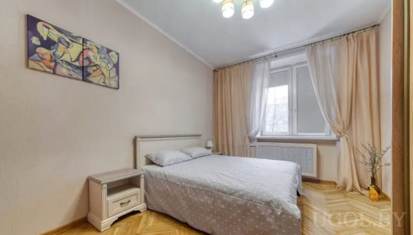 Уютная спальня с двуспальной кроватью, светлым покрывалом в горошек, прикроватной тумбой и яркой абстрактной картиной над изголовьем.