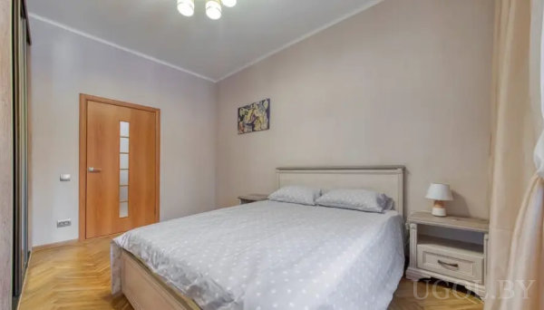 Минималистичная спальня с симметричными тумбами, светлым паркетом ёлочкой и деревянной дверью с матовыми вставками.