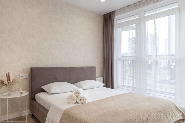 Современная спальня с кроватью в сером изголовье, белыми подушками, бежевым пледом и свернутыми полотенцами у больших окон с видом на город.