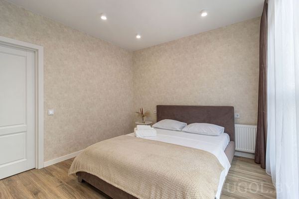 Минималистичная спальня с кроватью в коричневом изголовье, белым бельём, бежевым пледом и полотенцами, рядом — белая дверь и окно с тюлем.