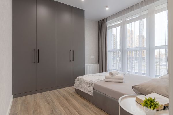 Современная спальня с серым шкафом, кроватью с белым бельём и свернутыми полотенцами, рядом — тумбочка с зелёным растением и панорамные окна.