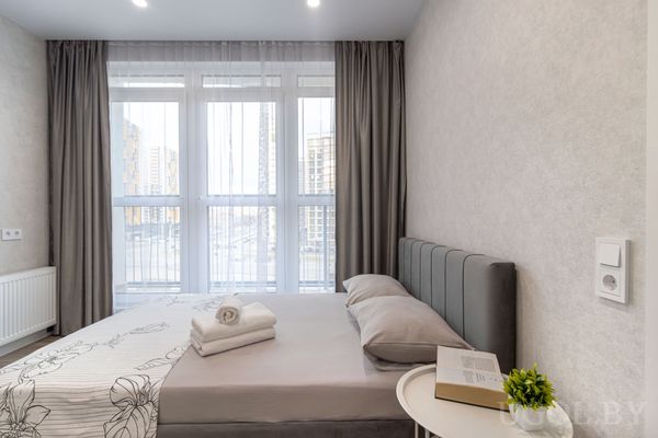 Уютная спальня с серым изголовьем, белыми подушками, бежевым пледом и книгой на круглой тумбочке у окна с видом на город.