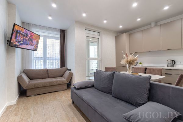 Современная гостиная с серым и коричневым диванами, телевизором на стене, обеденной зоной и большими окнами с видом на город.