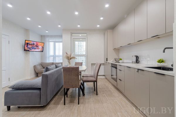 Открытая кухня с серыми шкафами, белой столешницей, круглым столом и мягкими стульями, рядом — диван и телевизор.