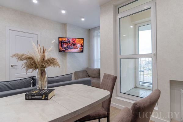 Светлая гостиная с круглым столом, серым диваном и телевизором с осенним изображением, рядом — дверь и выход на балкон.